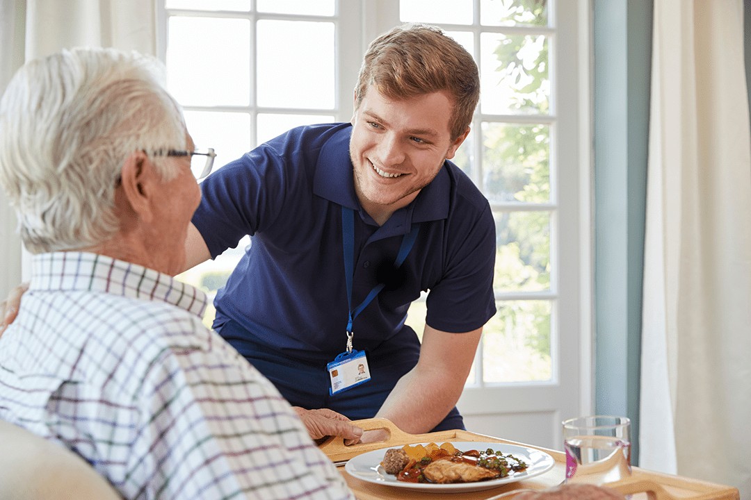 Male care worker serving dinner to a senior man at his home.