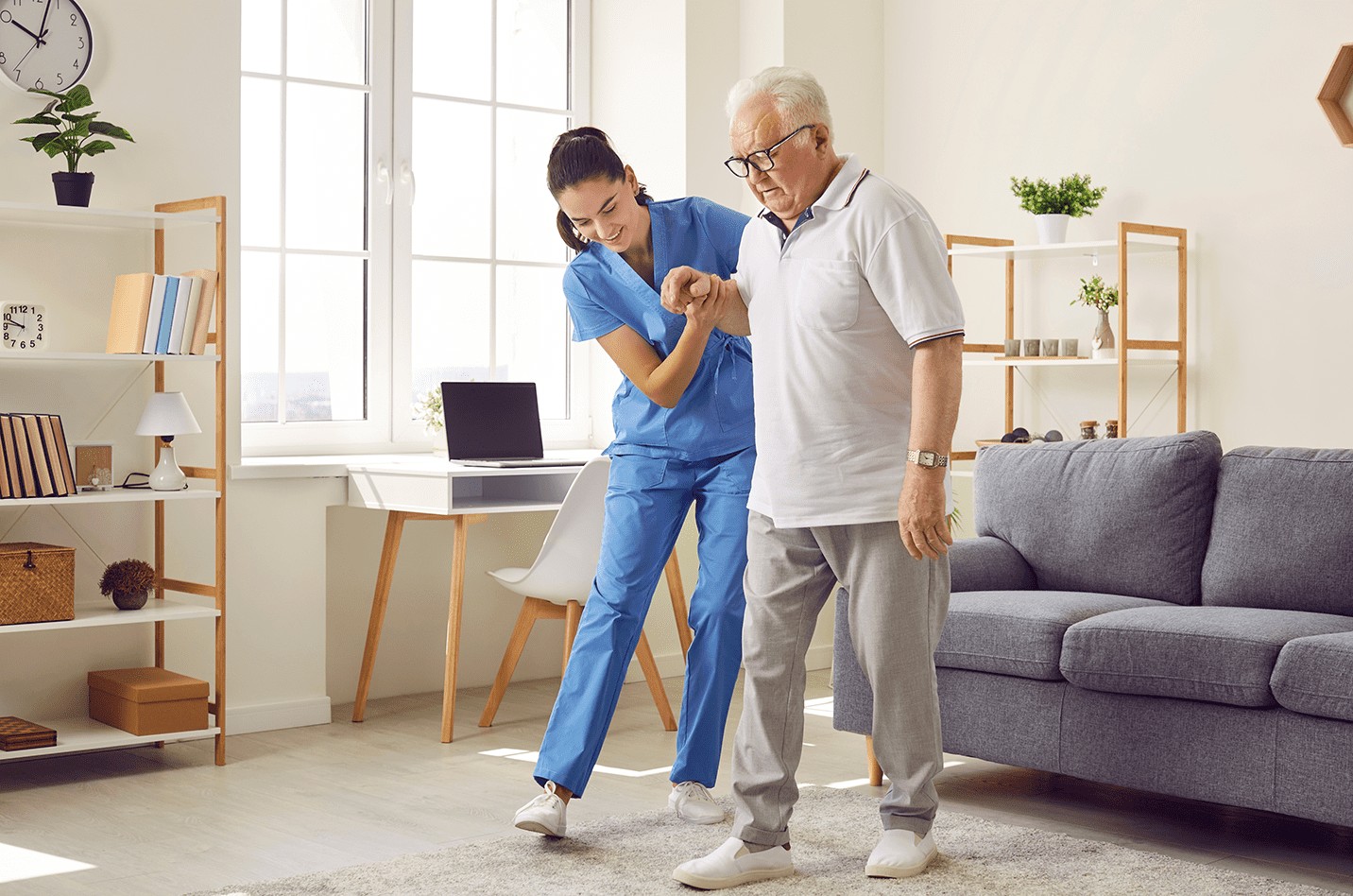 Young nurse helping elderly man walk in the room, holding his hand, supporting him. Home Health physical therapy treatment and rehabilitation after injury or stroke.