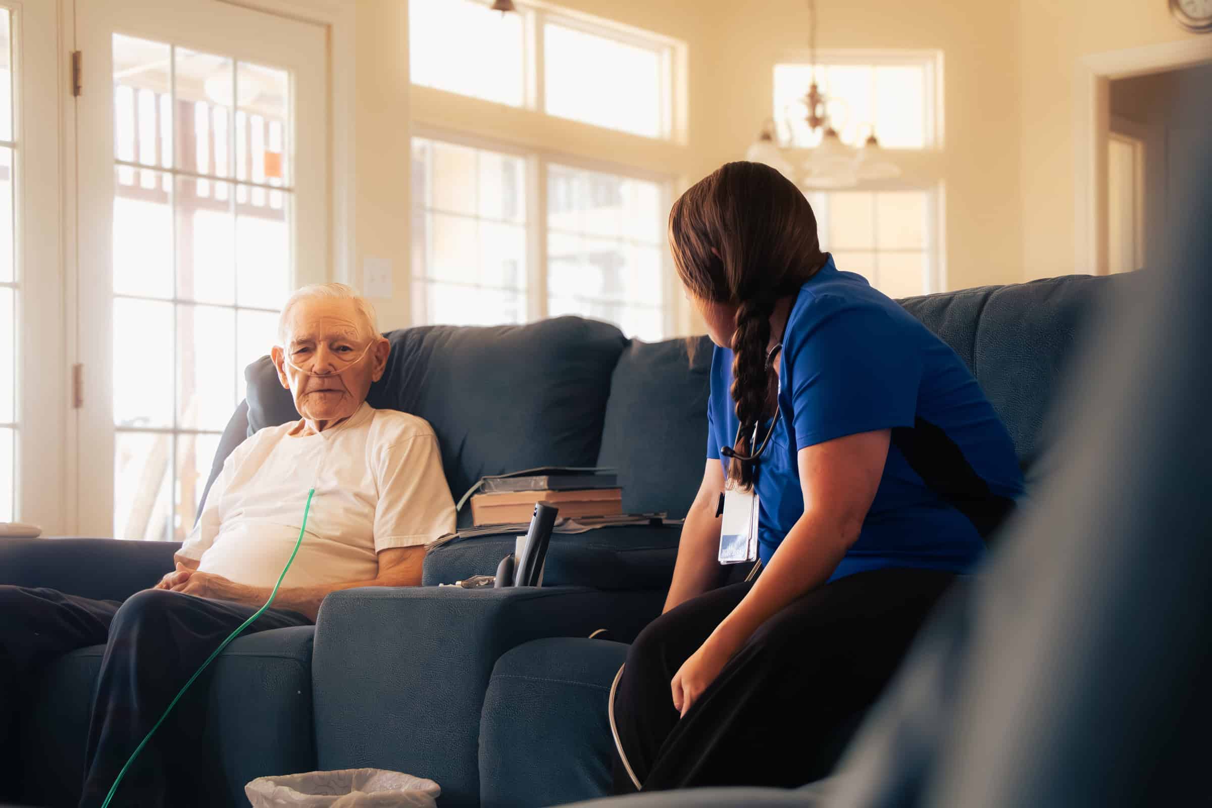 Hospice or home health clinician in blue scrubs sits beside an older adult patient using oxygen in a bright living room.